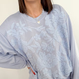 Person wearing a light gray sweatshirt with floral lace overlay on a plain background