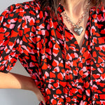 Person wearing a red and black patterned blouse with a heart necklace against a light background