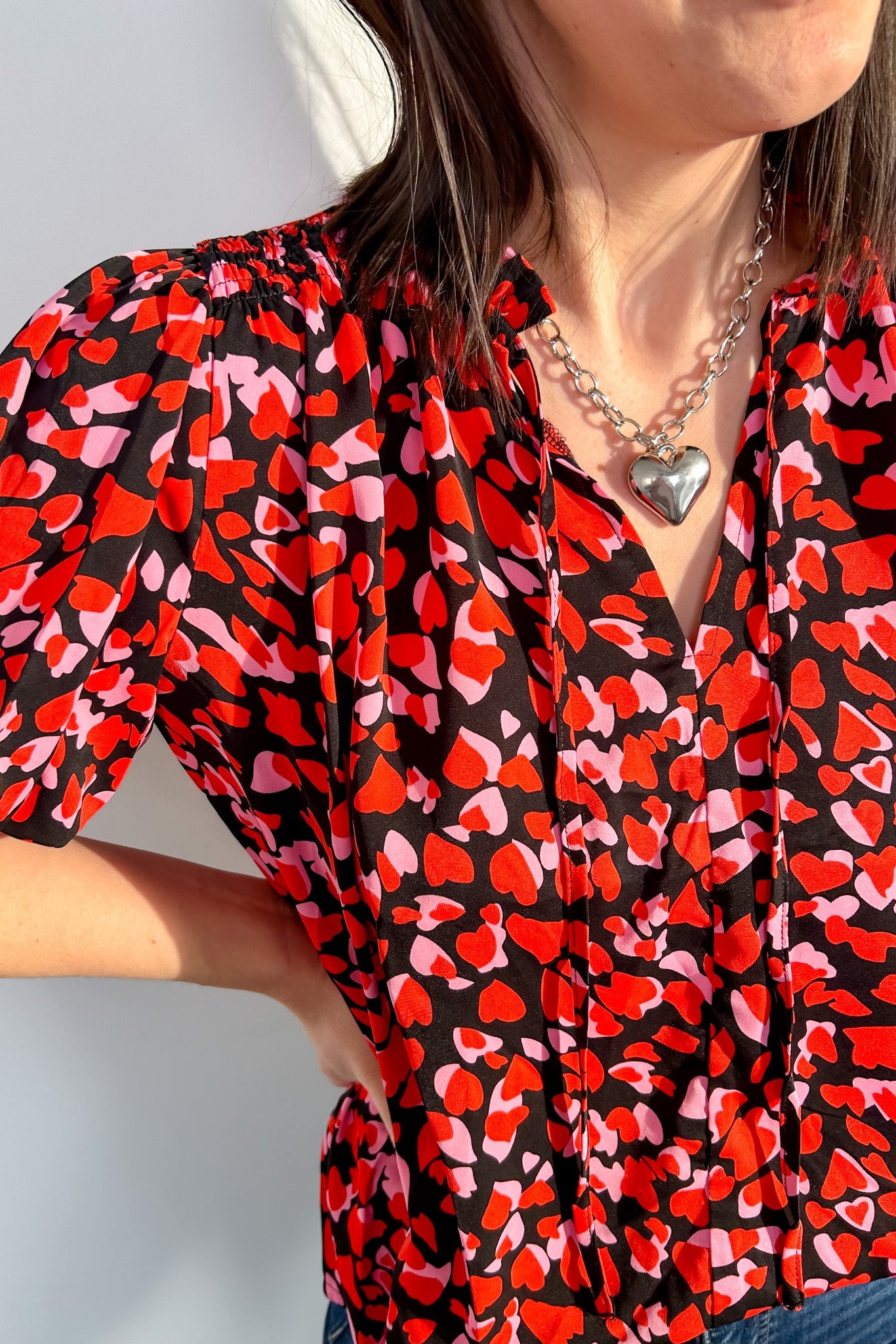 Person wearing a red and black patterned blouse with a heart necklace against a light background