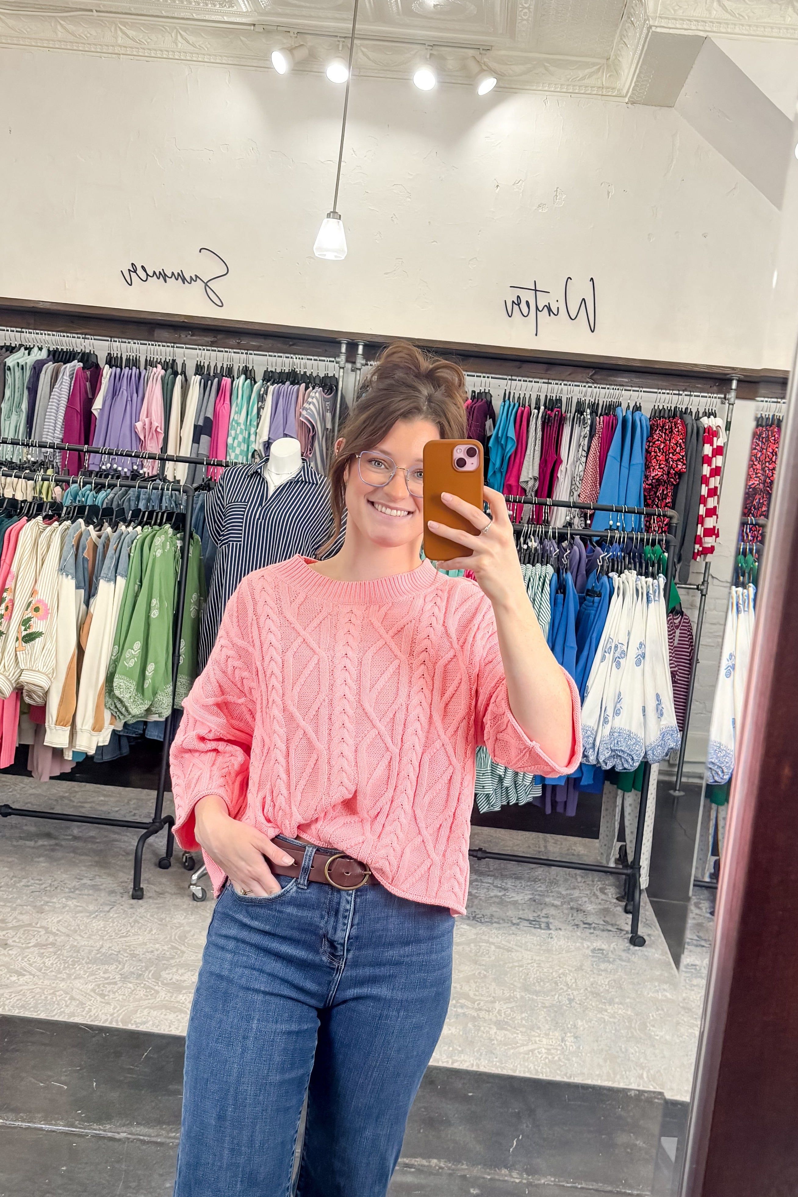 Person taking a mirror selfie wearing a pink sweater and blue jeans in a clothing store.
