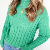 Person wearing a green ribbed sweater and blue jeans against a white background