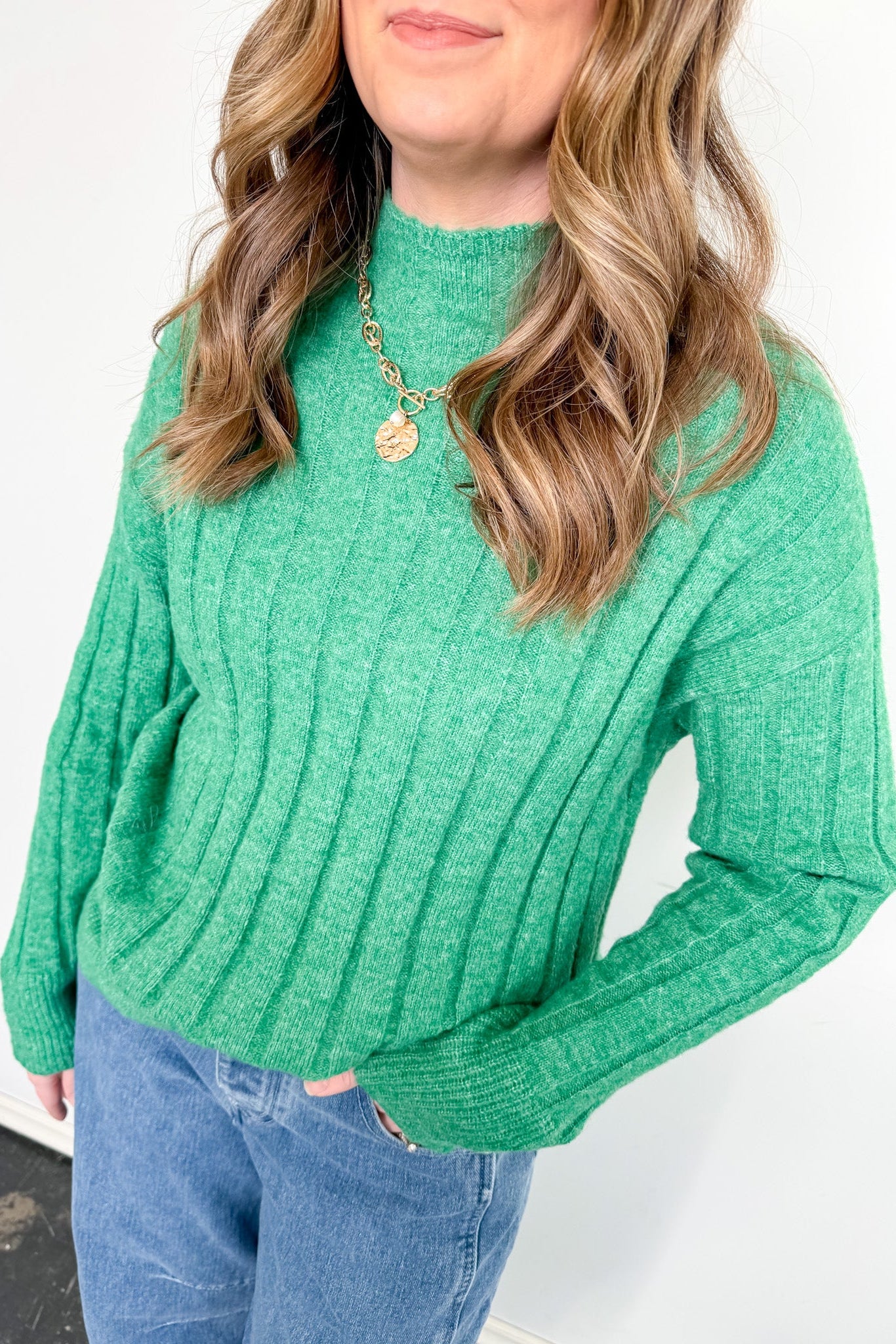 Person wearing a green ribbed sweater and blue jeans against a white background