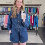 Woman wearing a denim romper that is collared with a tie waist and pockets.