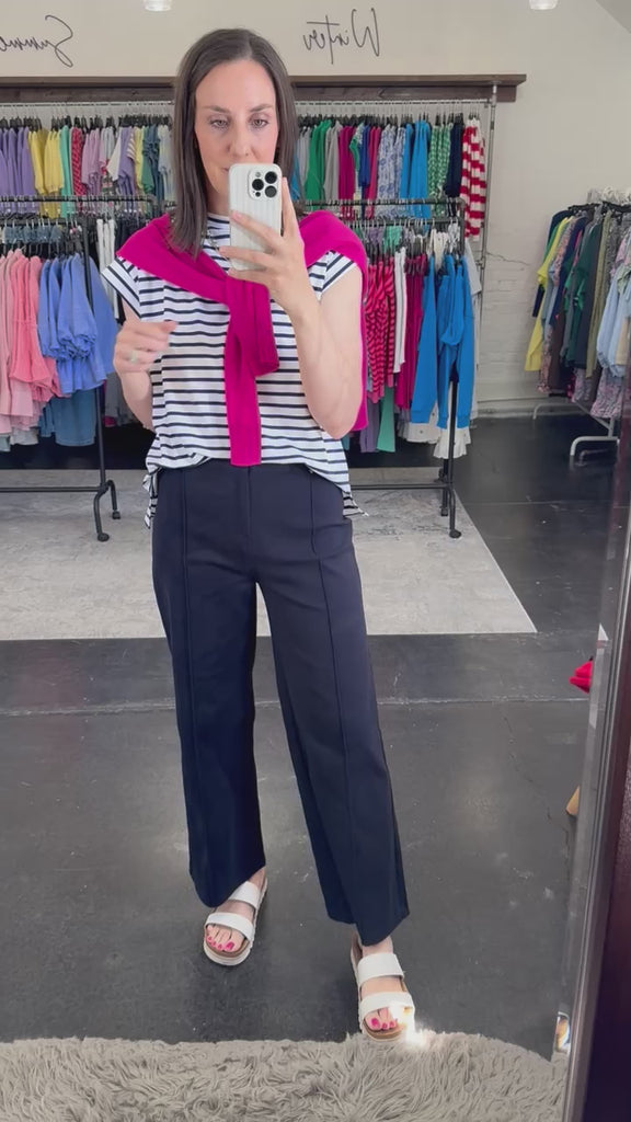 Woman wearing a striped blue and white top with a hot pink cardigan over her shoulders and navy pants. 